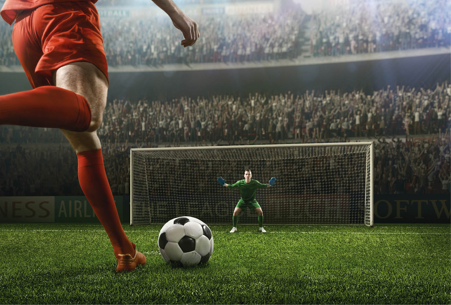 A male soccer player kicks a ball and soccer goalie jumping in motion for a ball while defending his gates on wide angle panoramic image of a outdoor soccer stadium or arena full of spectators under a sunny sky. The image has depth of field with the focus on the foreground part of the pitch. With intentional lensflares. Players are wearing unbranded soccer uniform.