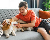 Happy teenager with down syndrome relaxing on a comfortable sofa at home and petting his welsh corgi pembroke dog. Friendship, animal concept