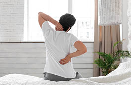 Unrecognizable guy suffering from back pain in bedroom, waking up in morning, sitting on bed and touching his neck and lower back with both hands, wearing pajamas, copy space, back view