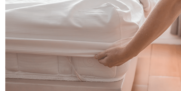 A woman makes a bed in a bedroom, securing a white fitted sheet to a polyurethane mattress.