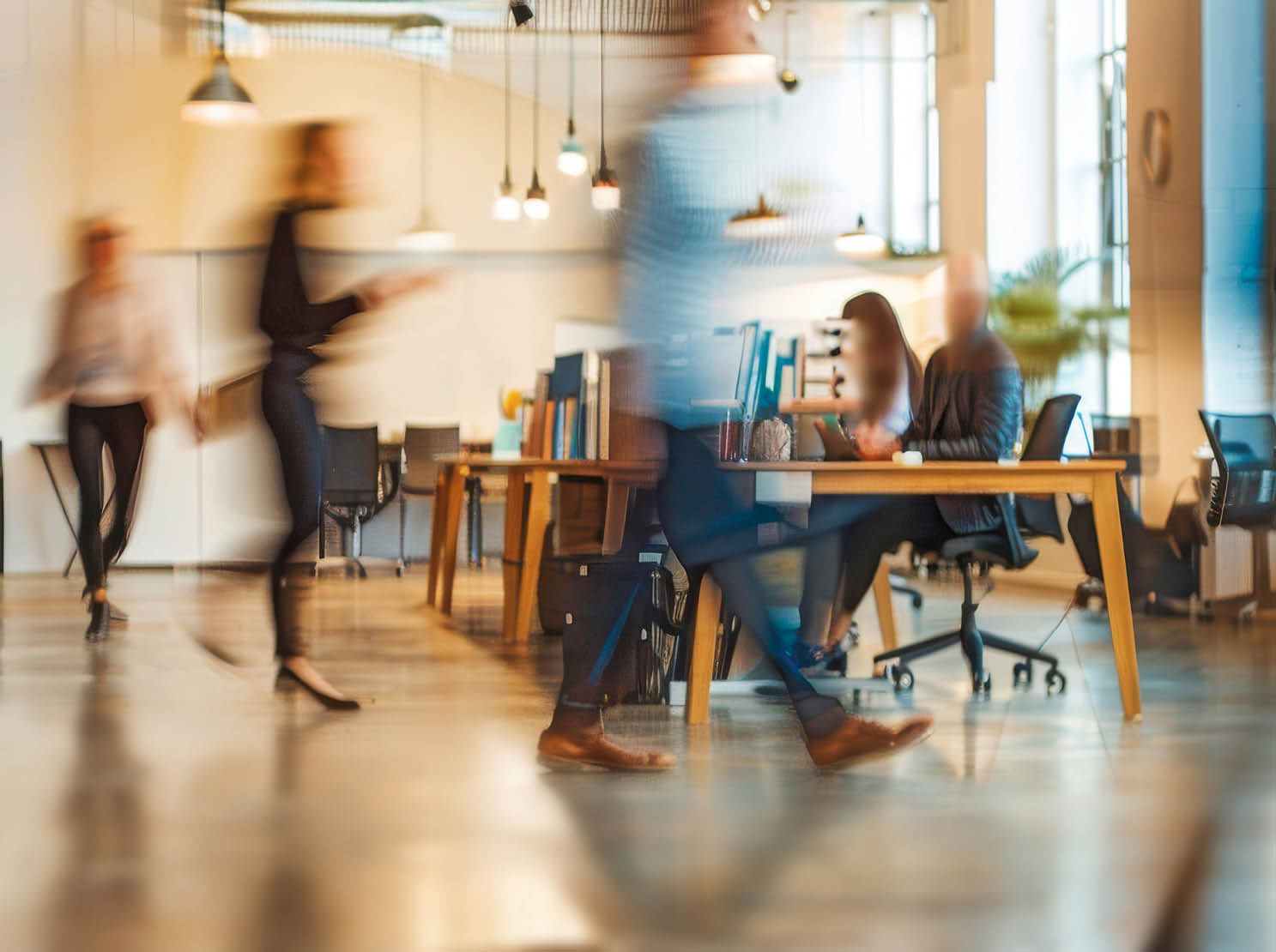This photo captures a modern office space with several blurred figures of business professionals going about their work. The image conveys a sense of movement and activity within the workplace.