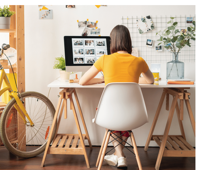 Rear view of young brunette female sitting on white chair in front of computer monitor while looking throguh collection of photos
