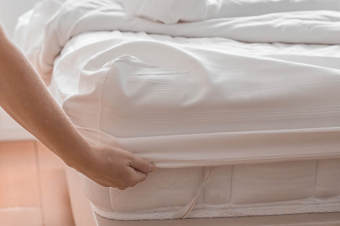 A woman makes a bed in a bedroom, securing a white fitted sheet to a polyurethane mattress.