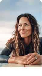A woman surfer lying on her surfboard in the ocean, sun shining behind, looking content as she glides over the water. 