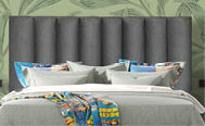 A neatly made bed with a tall, dark gray upholstered headboard, light gray bedding, colorful pillows, and a vibrant patterned blanket draped across the foot of the bed against a green leafy wallpaper background. Description generated by AI