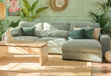 A large, light grey couch is placed in a living room with a window behind it. The couch is positioned in front of a potted plant and a vase. AI generated content