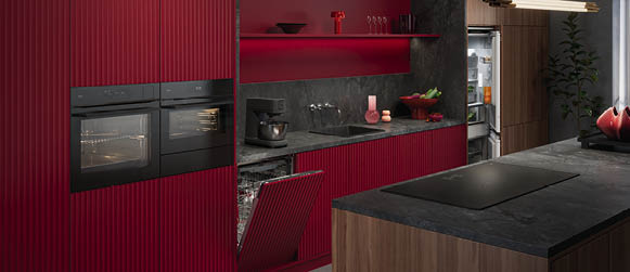 A kitchen with red cabinets and a black countertop. A stove and oven are visible, along with a sink and a refrigerator. There are several bottles and a bowl on the countertop. Conte do gerado por IA