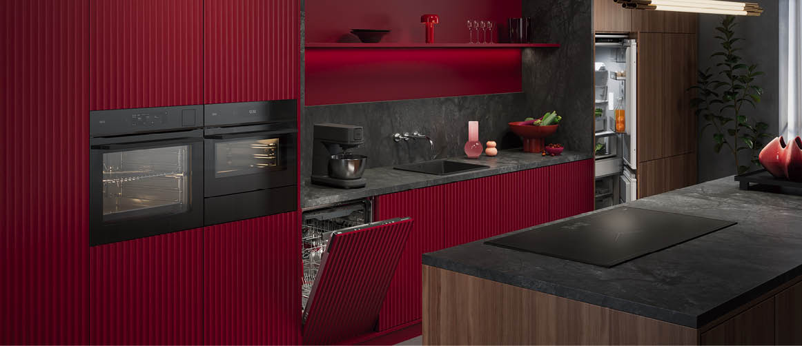 A kitchen with red cabinets and a black countertop. A stove and oven are visible, along with a sink and a refrigerator. There are several bottles and a bowl on the countertop. Conte do gerado por IA