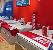 A showroom displays several mattresses with red and white bedding and promotional materials against a red and blue wall. Description generated by AI
