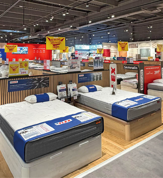 A brightly lit mattress store displays multiple mattresses on wooden platforms with promotional signs hanging from the ceiling and placed on the mattresses. Description generated by AI