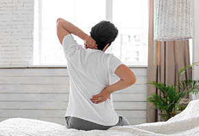Unrecognizable guy suffering from back pain in bedroom, waking up in morning, sitting on bed and touching his neck and lower back with both hands, wearing pajamas, copy space, back view