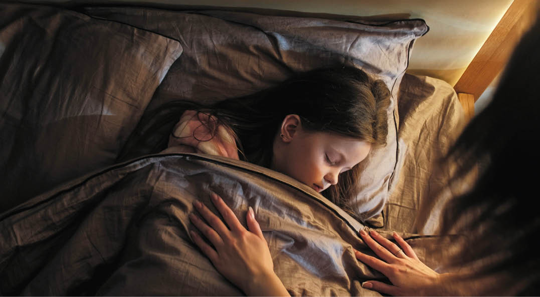 Sleeping child mother care. Night rest. Woman covering peaceful calm daughter with blanket lying in bed at late night in dark home bedroom.