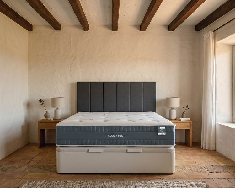 A neatly made bed with a gray upholstered headboard is centered between two wooden nightstands with matching lamps and small decorative items in a rustic room with exposed wooden ceiling beams and a large window with white curtains. Description generated by AI