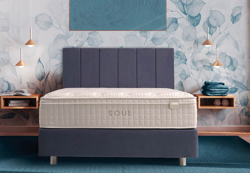 A modern bedroom features a neatly made bed with a quilted mattress labeled “SOUL," flanked by wooden nightstands holding folded towels and books, set against a floral wallpaper and illuminated by hanging pendant lights. Description generated by AI