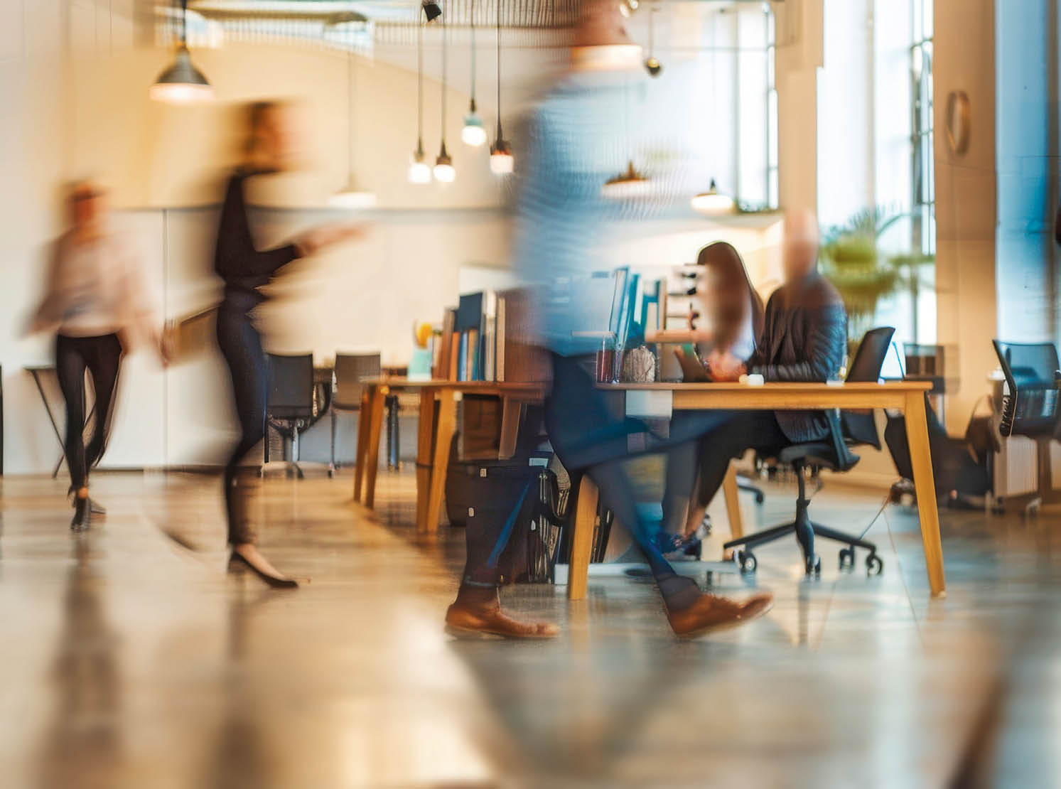 This photo captures a modern office space with several blurred figures of business professionals going about their work. The image conveys a sense of movement and activity within the workplace.