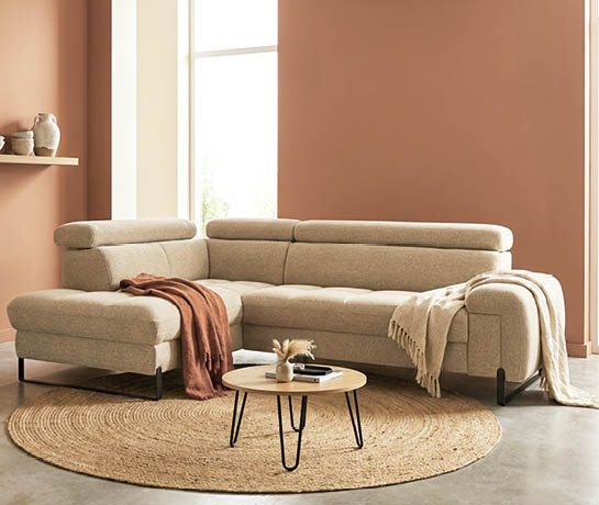 A large, tan couch is placed in front of a window. The couch is positioned in front of a wall, and there is a rug underneath it. A vase is also present in the scene. AI generated content